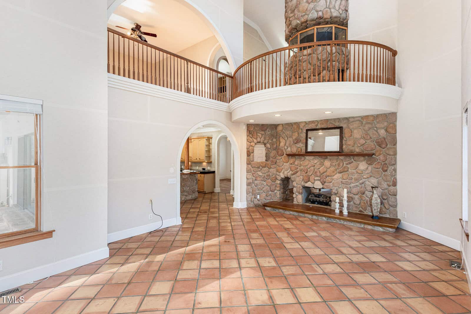A spacious living room with terracotta tile flooring, a stone fireplace, vaulted ceilings, a curved upstairs balcony, and an archway leading to a kitchen area. Sunlight streams in from windows on the left.