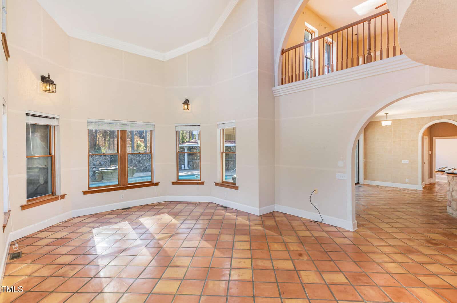 A spacious interior room with high ceilings, terracotta tile flooring, cream-colored walls, large windows, an arched doorway, and an open upstairs balcony overlooking the area below.