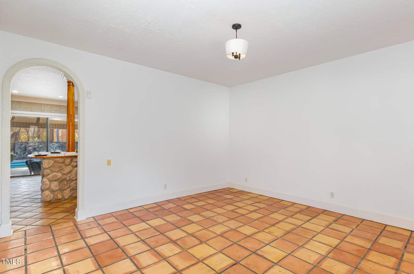 Empty room with terracotta tile floor, white walls, and a modern ceiling light. An arched doorway leads to another area with a stone accent and a view of a swimming pool outside.