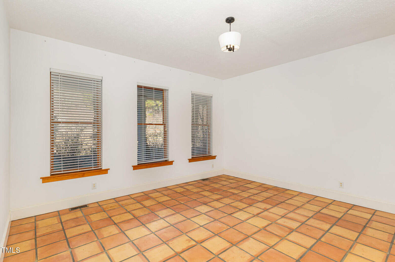 A bright, empty room with white walls, three windows with blinds, a ceiling light, and a floor of terracotta tiles.