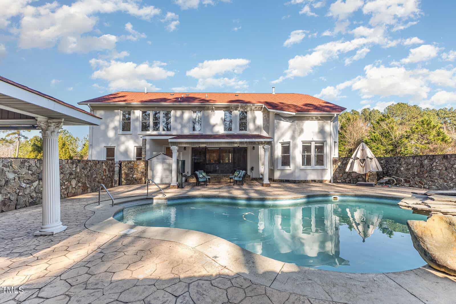 A large two-story white house with a red roof stands behind a backyard with a freeform swimming pool, poolside lounge chairs, an umbrella, and a textured patio under a partly cloudy sky.