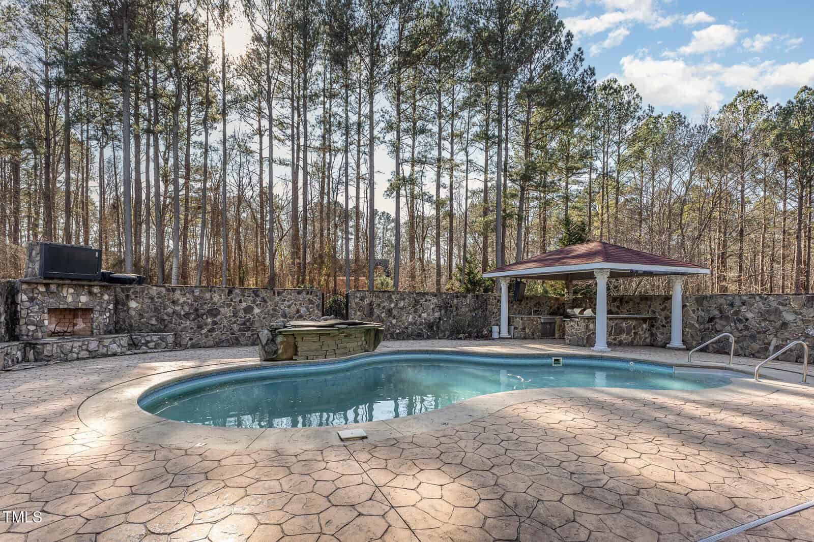 A backyard pool with curved edges set in a stamped concrete patio, surrounded by a stone wall, with a covered gazebo, a hot tub, and a fireplace; tall trees and blue sky in the background.