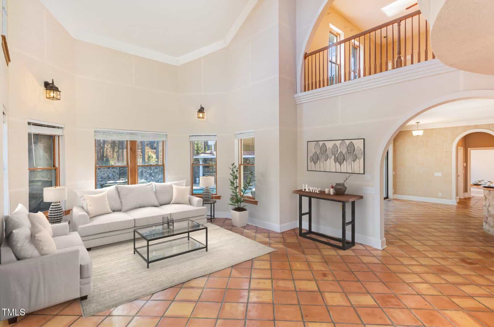 Spacious living room with terracotta tile floors, white walls, a large gray sectional sofa, glass coffee table, potted plant, console table with decor, and an open second-story balcony overlooking the area.