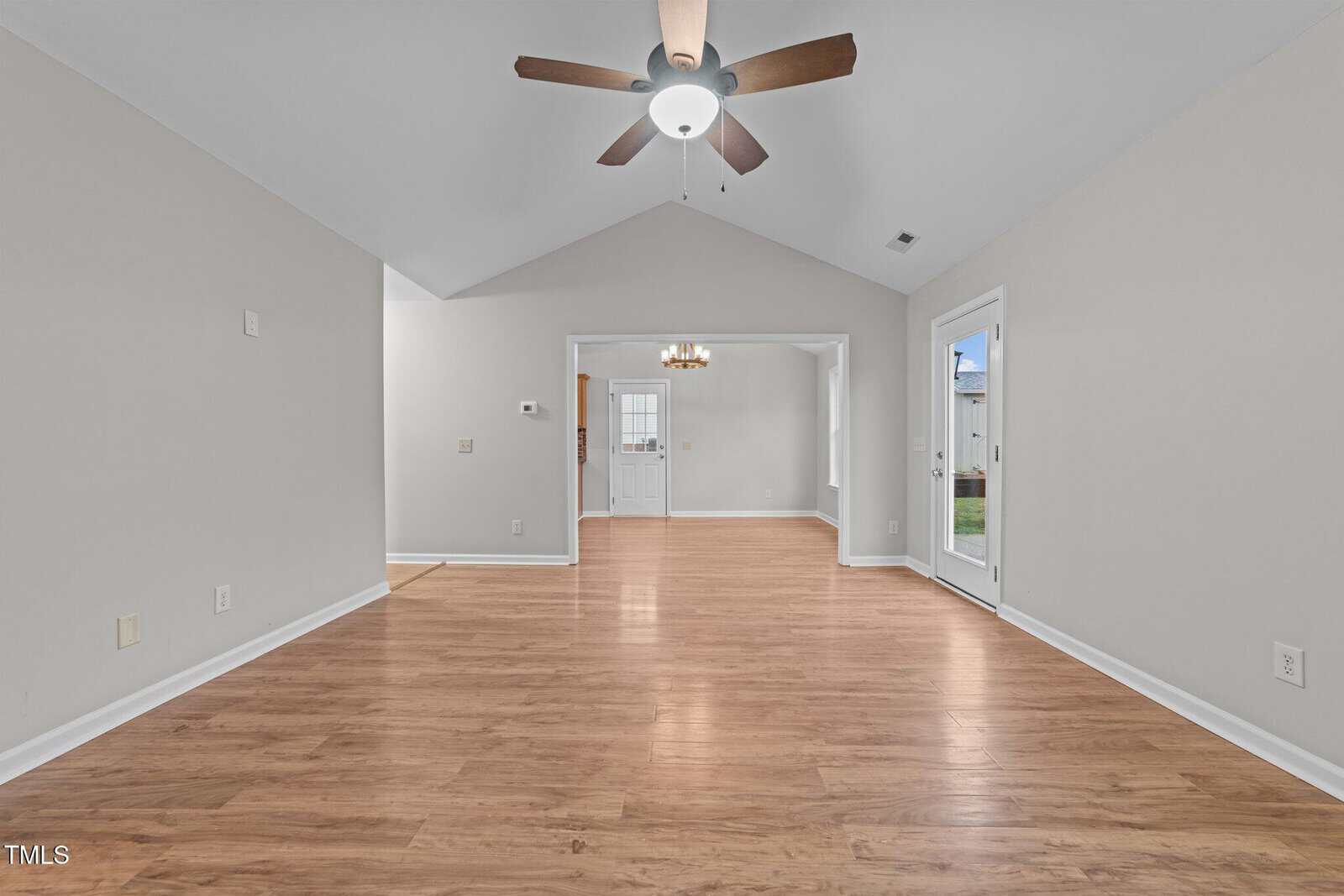 A spacious, empty living room with light wood flooring, beige walls, a ceiling fan with a light, and white trim. There are doors leading outside and into an adjacent dining area and entryway.