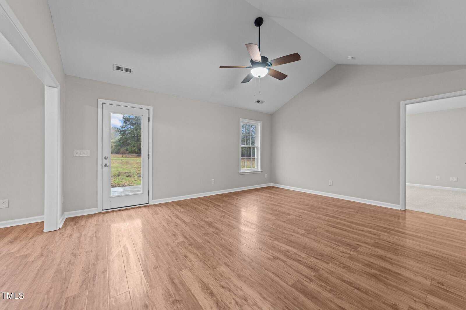 A spacious, empty room with light brown wood flooring, light gray walls, a ceiling fan with lights, a window, a glass door leading outside, and an open doorway to another carpeted room.