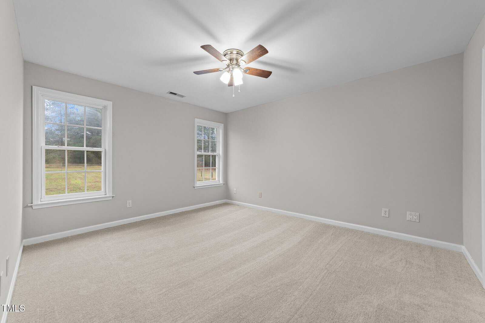 A vacant room with beige carpet, light gray walls, two large windows, a ceiling fan with lights, and electrical outlets on the walls.
