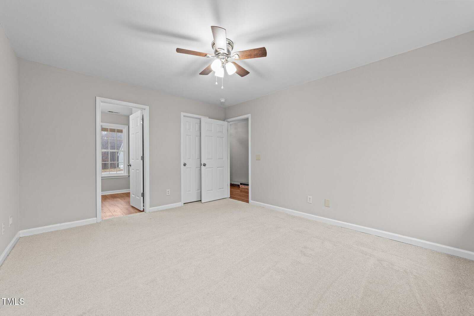 Empty bedroom with beige carpet, light gray walls, ceiling fan with lights, and three white doors; one door leads to a bathroom, another to a hallway, and one to a closet. Natural light comes from an adjacent window.