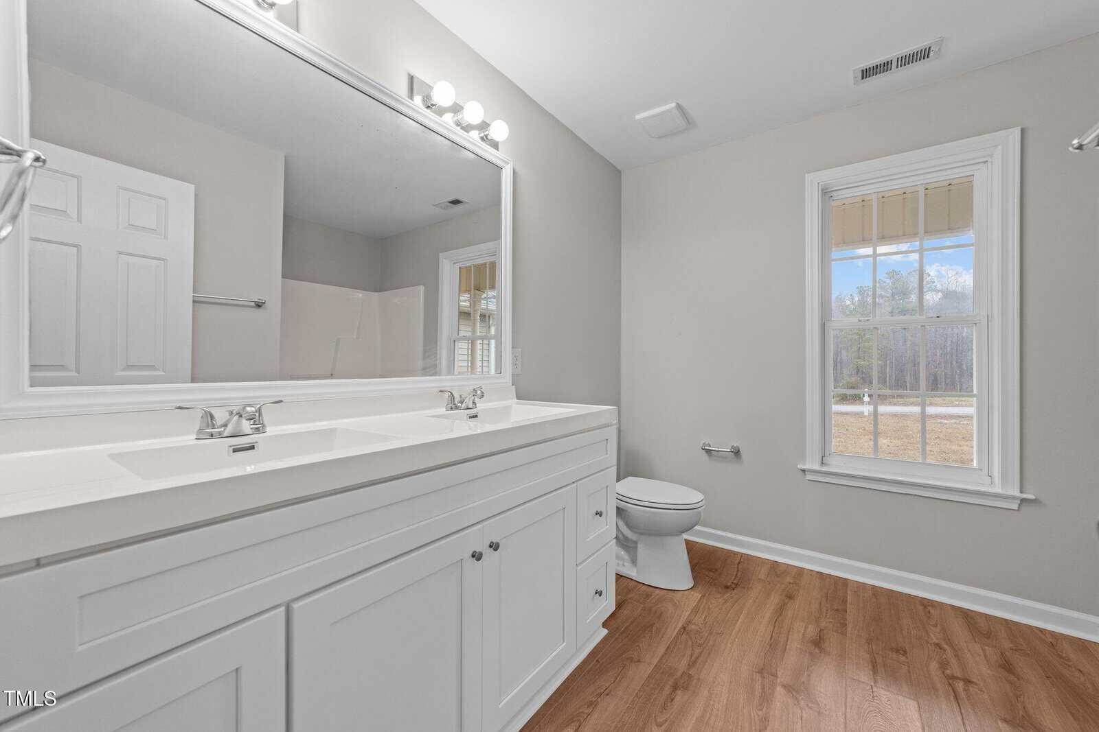 Bright bathroom with double sinks, large mirror, toilet, white cabinetry, and wood-style flooring. There’s a window letting in natural light and beige walls with modern fixtures.