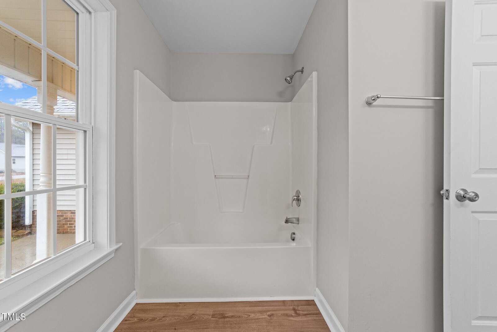 White bathtub and shower with built-in shelves, silver fixtures, and a towel bar next to a closed white door. Large window lets in natural light and shows part of the house exterior. Wood flooring is visible.