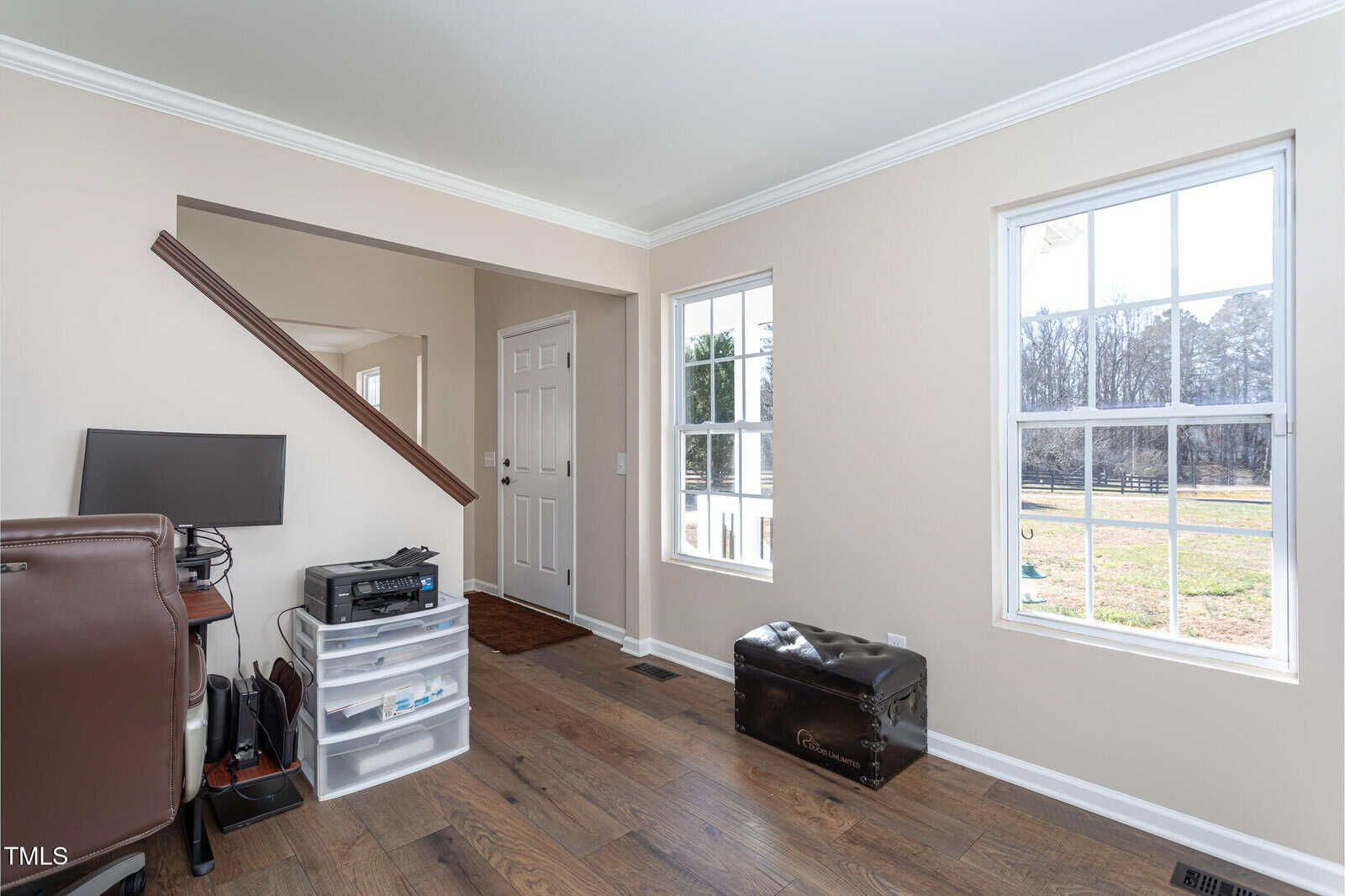 A home office area with a computer desk, office chair, plastic drawer unit, and a trunk near two windows. The space features light-colored walls, wood flooring, and an entryway with a white door.