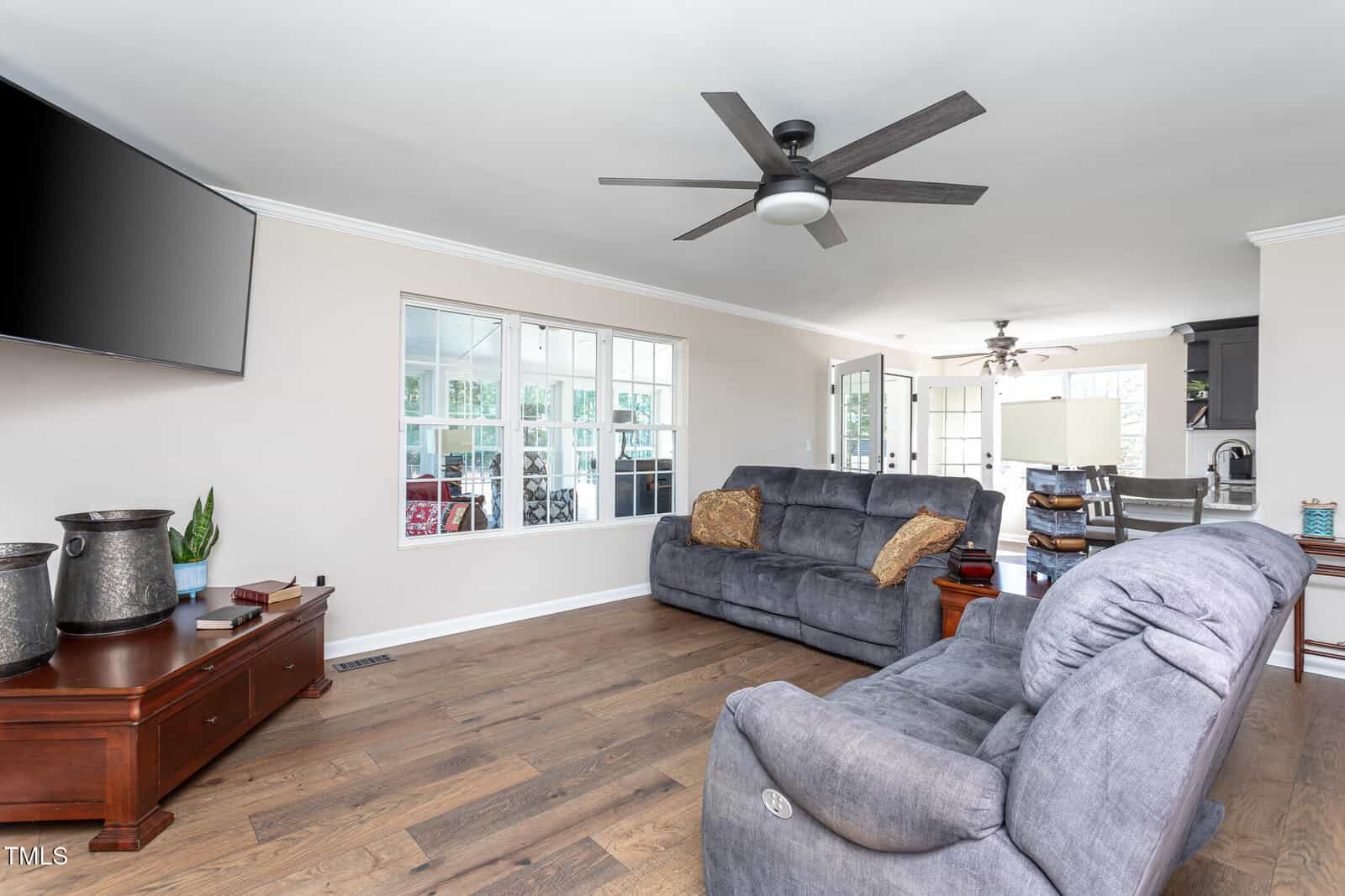 A living room with wood flooring, two gray recliner sofas, a ceiling fan, a wall-mounted TV, and a wooden coffee table; kitchen and dining area visible in the background.
