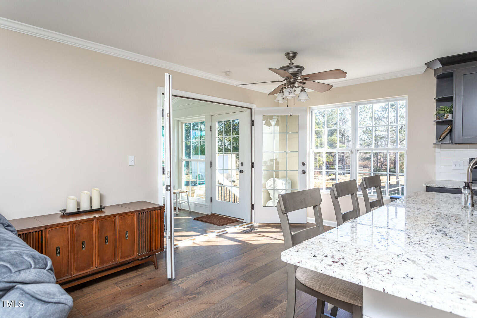 A bright kitchen area with granite countertops, wooden chairs, and a ceiling fan. An open door leads to a sunroom with large windows that let in natural light. A wooden sideboard with candles is against the wall.