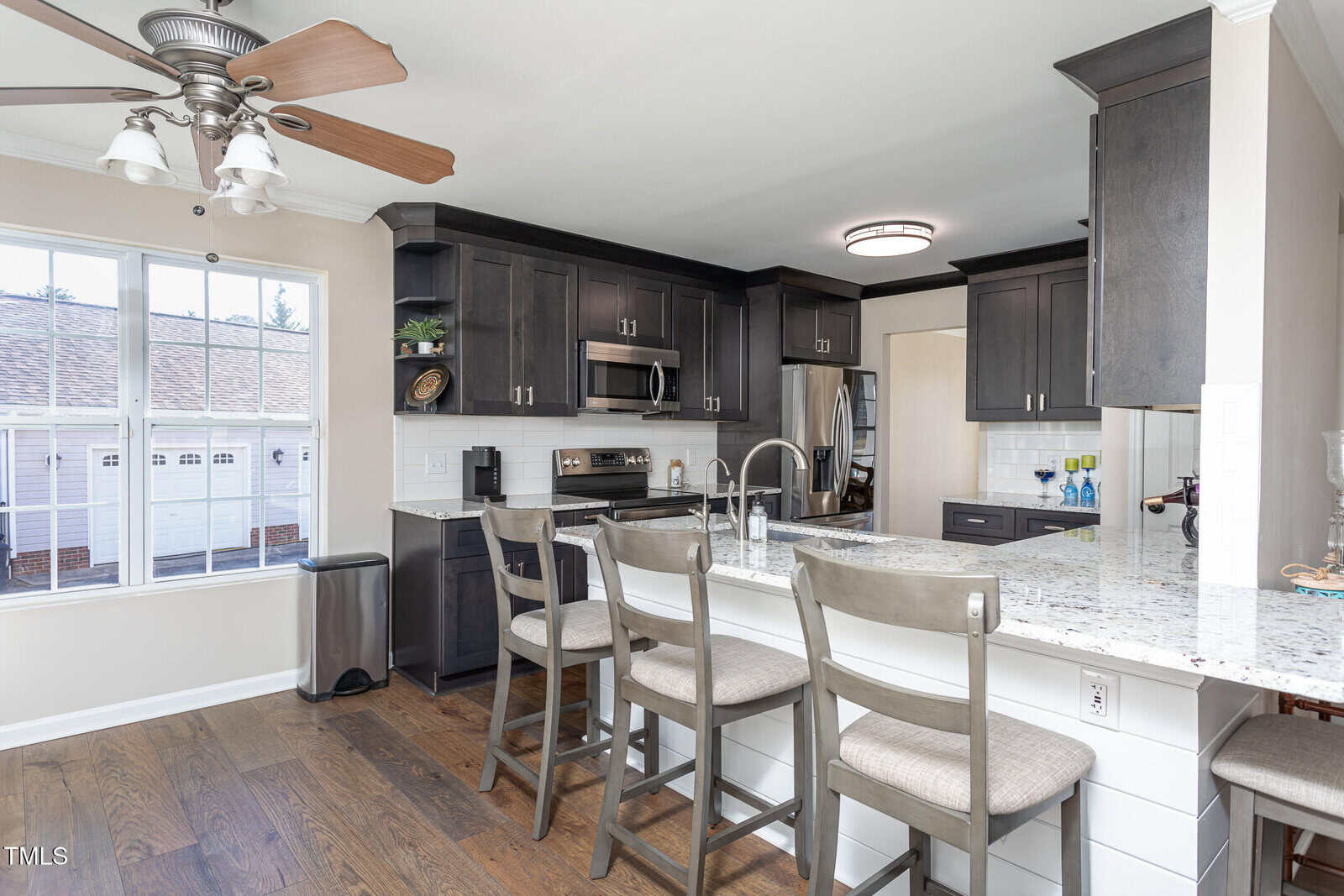 Modern kitchen with dark cabinets, stainless steel appliances, a large granite island with four gray barstools, wood floors, and a ceiling fan. Large window lets in natural light.