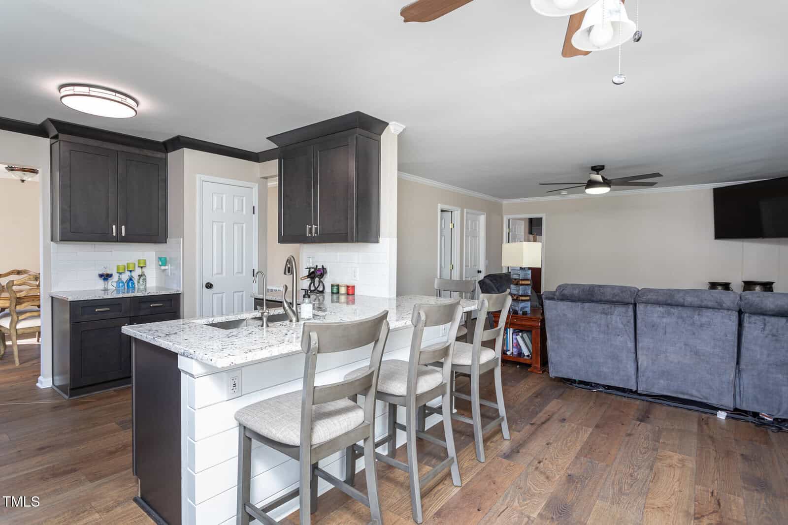 Modern kitchen with dark cabinets, white countertops, three gray barstools at an island, wood flooring, and an open view to a living area with gray sofas and ceiling fans. Dining room visible through a doorway.