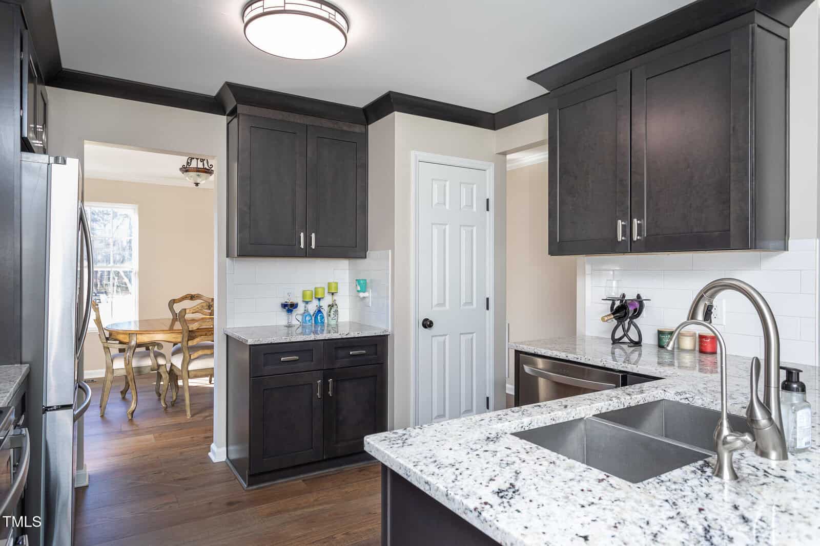 Modern kitchen with dark cabinets, white granite countertops, stainless steel sink, and white subway tile backsplash. A dining area with wooden chairs and table is visible through the doorway in the background.