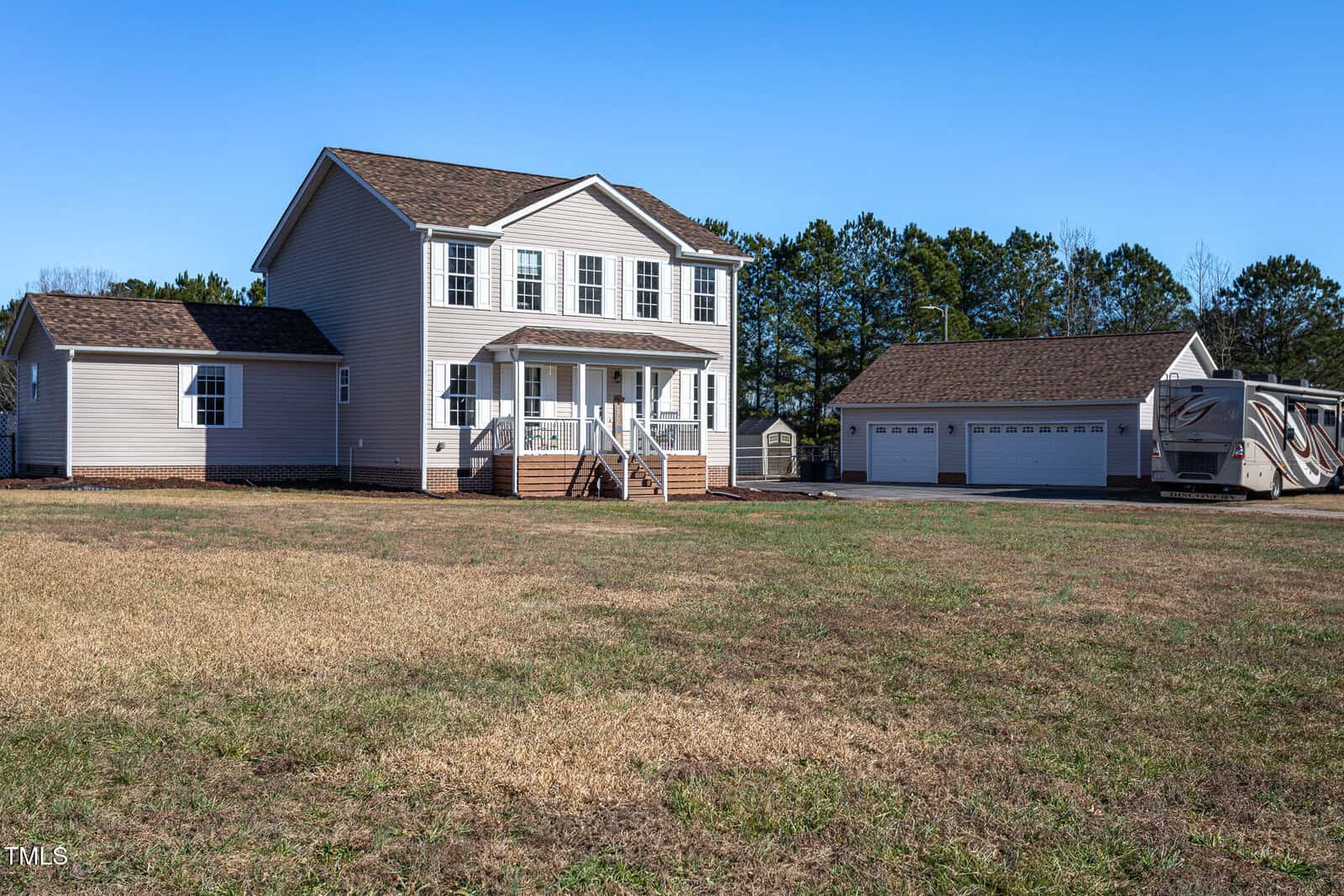 Two-story beige house with a covered front porch, detached three-car garage, and an RV parked nearby. The yard is spacious with patchy grass, and there are trees in the background under a clear blue sky.