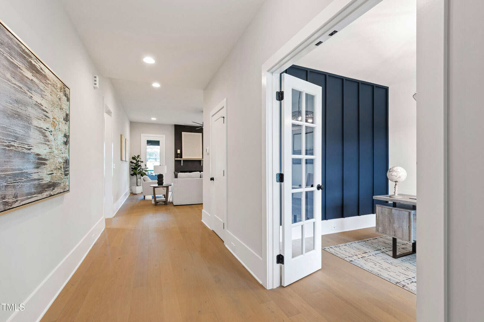 A hallway with light wood floors and white walls leads to a living area. On the right, French doors open to a room with dark blue paneled walls, a desk, and a globe. Contemporary decor is visible throughout.