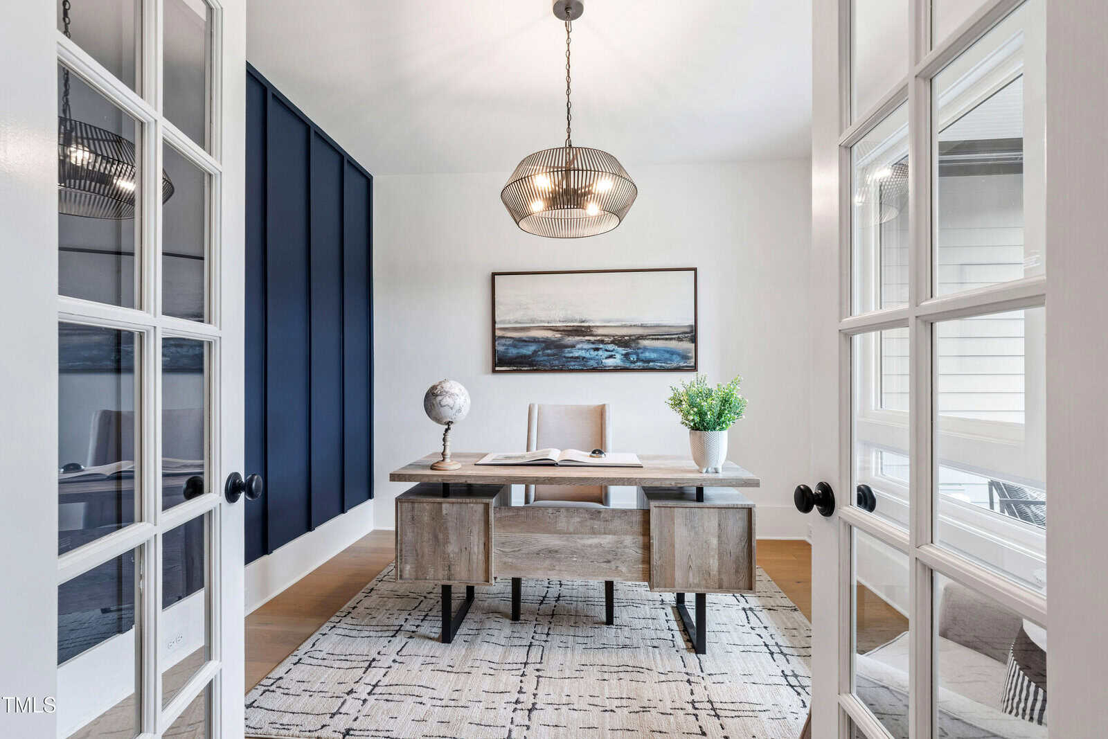 A modern home office with a wooden desk, chair, globe, potted plant, and artwork on the back wall. Double glass doors open into the room, which has a geometric rug and a contemporary ceiling light fixture.
