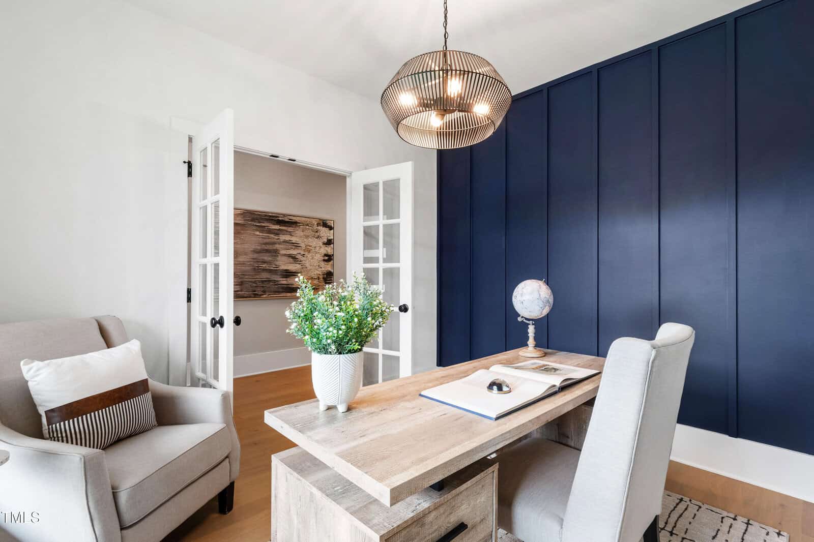 A modern home office with a wooden desk, gray upholstered chair, globe, open book, potted plant, and a blue accent wall. French doors open to another room with artwork visible on the wall.