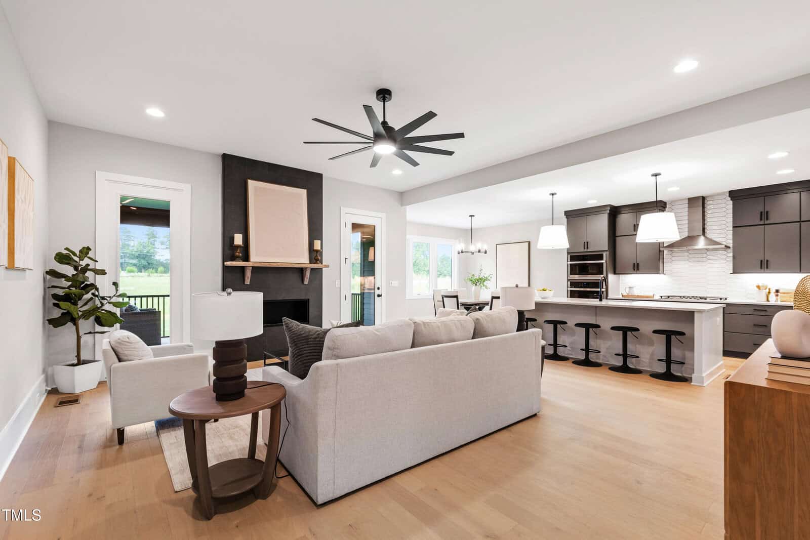 Modern open-concept living room and kitchen with neutral colors, light wood floors, gray sofa, round side table, black accent fireplace, barstools at kitchen island, pendant lights, and large windows letting in natural light.