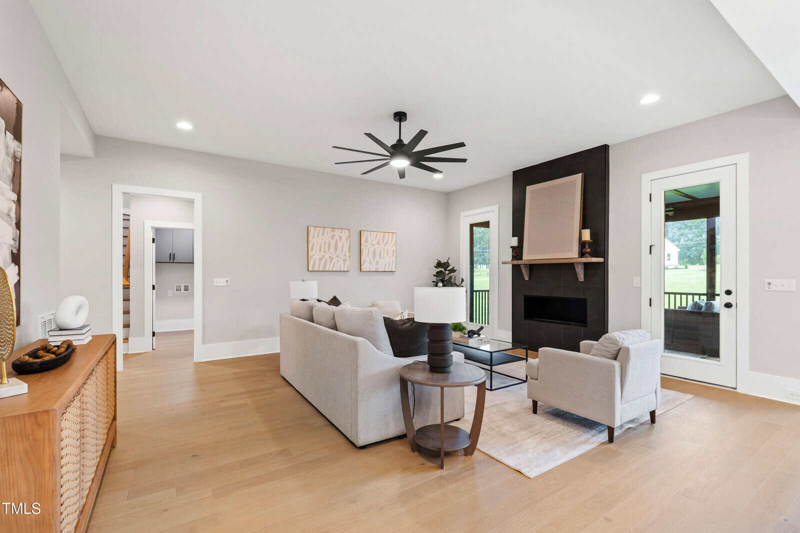 Modern living room with light wood floors, beige walls, gray sofas, a black fireplace with a wooden mantle, and large windows letting in natural light. Decor includes abstract wall art and neutral-toned accessories.