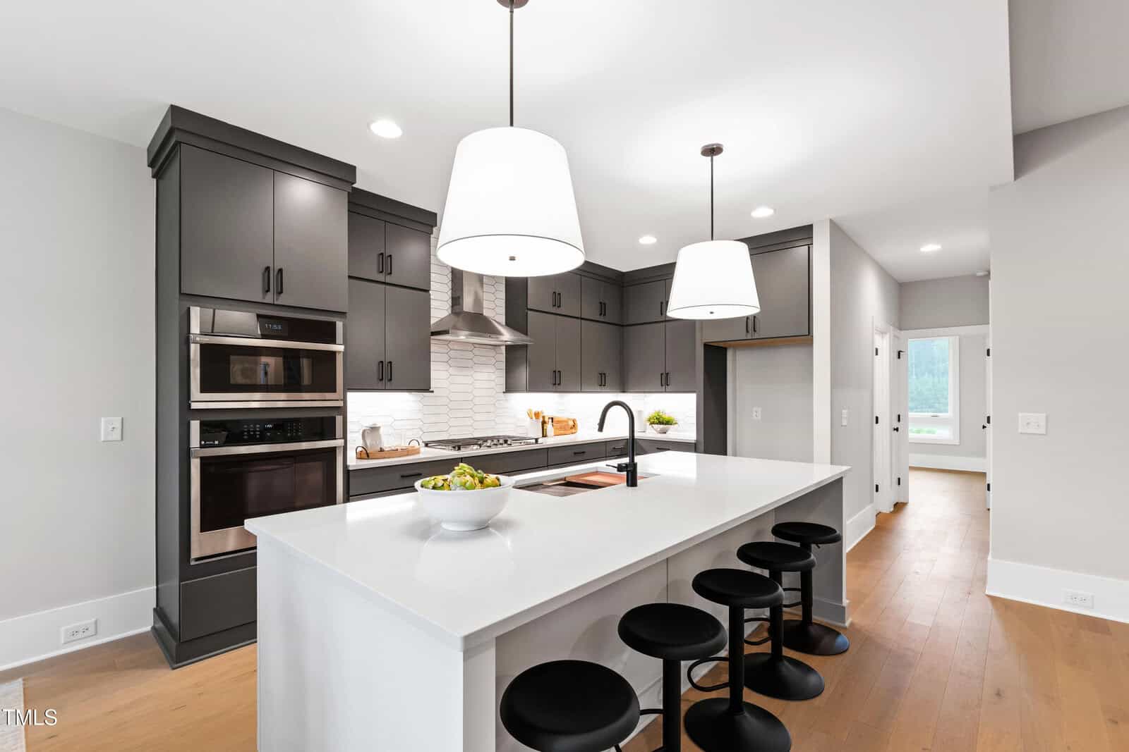 A modern kitchen with gray cabinets, stainless steel appliances, a white island with a built-in sink, four black stools, white subway tile backsplash, and two large pendant lights hanging above the island.