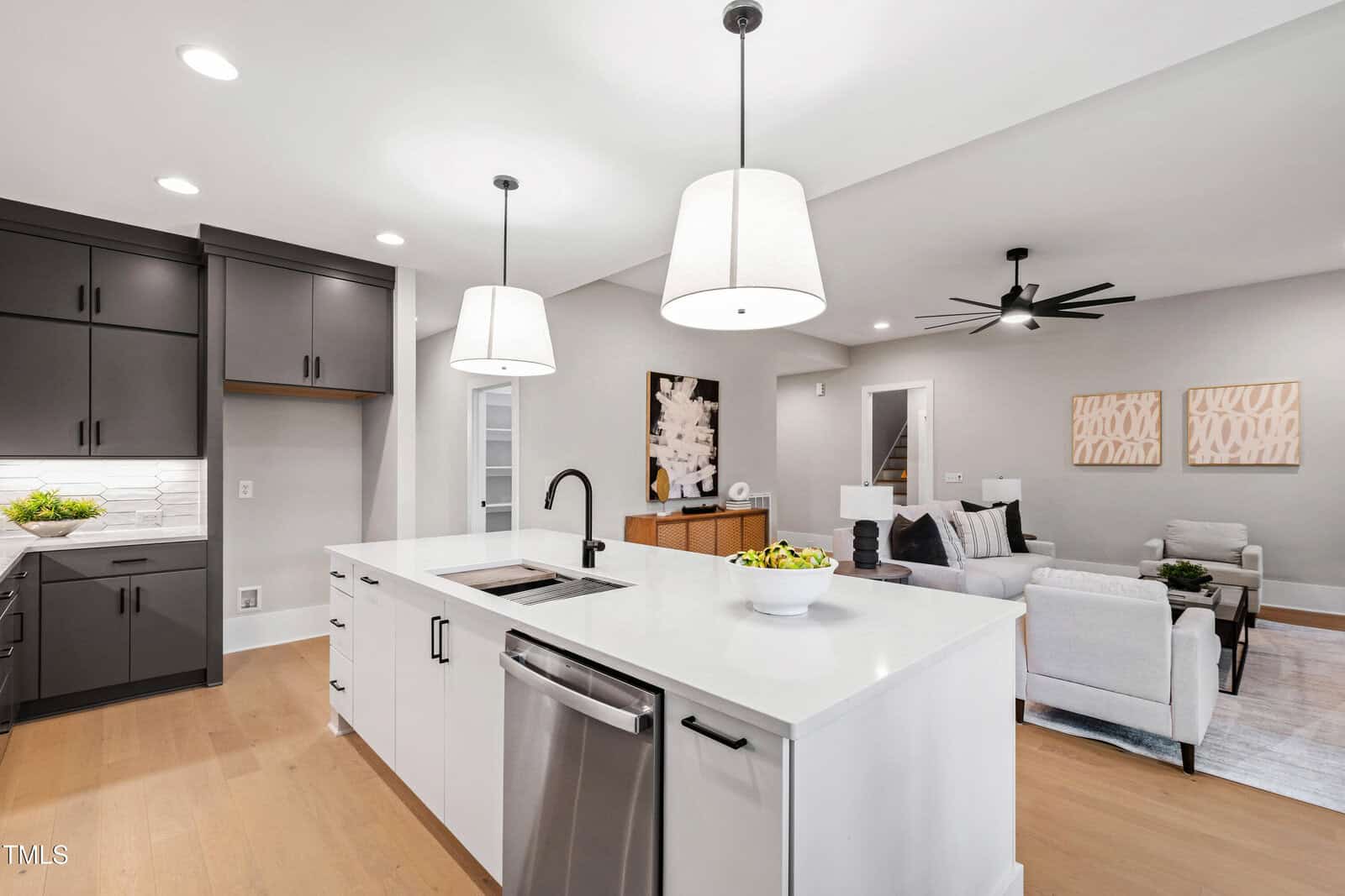 A modern open-concept kitchen and living room with white walls, light wood floors, a white island with a sink, stainless steel appliances, black cabinets, pendant lights, and neutral-colored furniture in the living area.