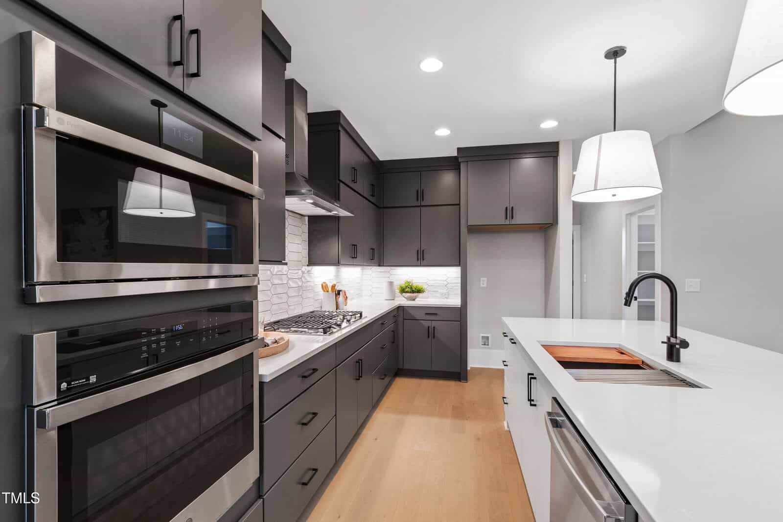Modern kitchen with dark gray cabinets, stainless steel appliances, built-in oven and microwave, white tile backsplash, light wood flooring, and a white countertop with a sink and pendant lights overhead.