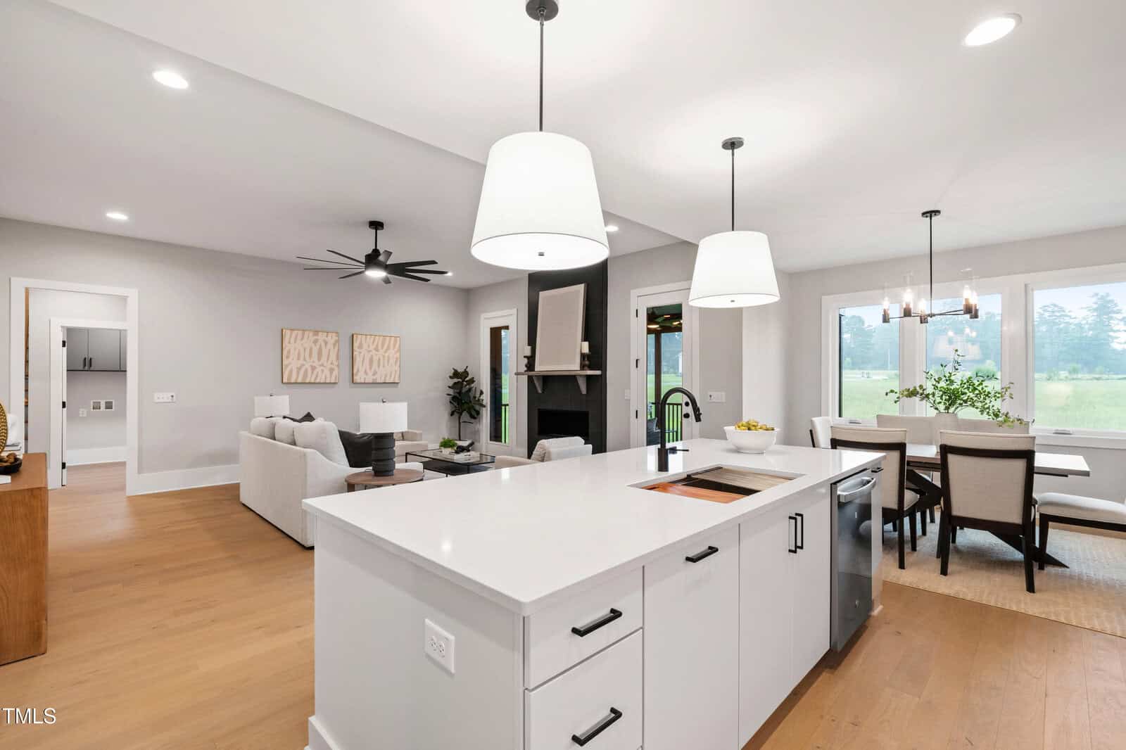 A modern open-concept kitchen and living space features a white island with a sink, pendant lights, a dining table by large windows, a ceiling fan, and light wood floors.