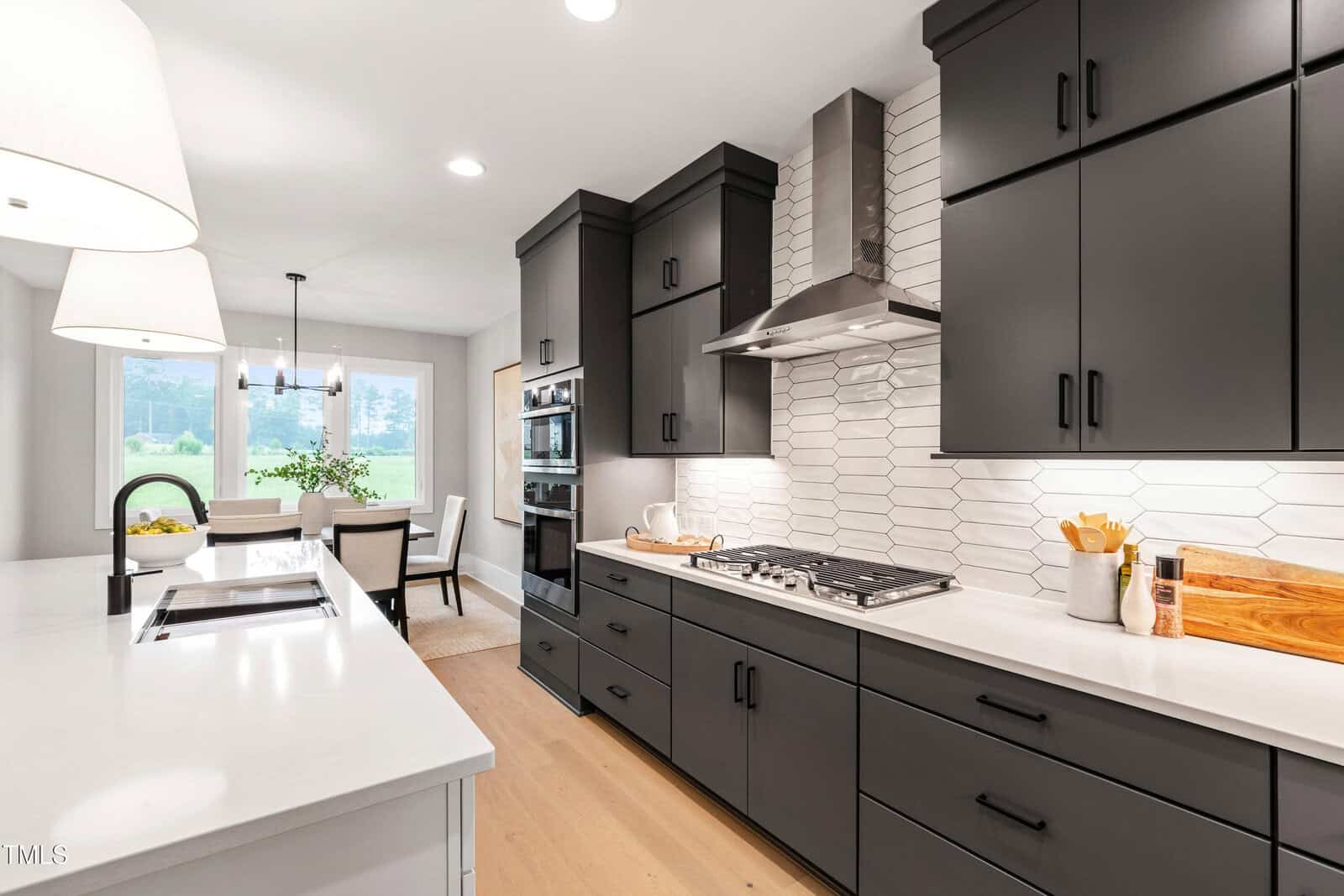 Modern kitchen with matte black cabinets, white countertops, a stainless steel hood over a gas stove, white hexagonal tile backsplash, light wood floors, and a dining area visible in the background.