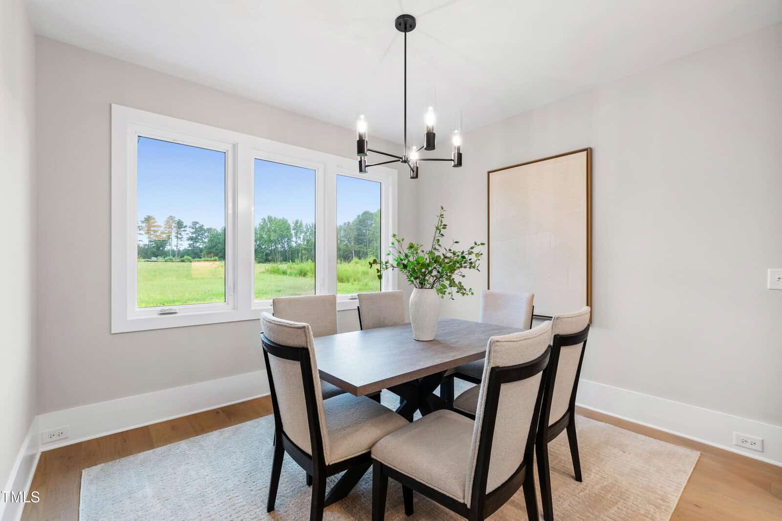 A bright dining room features a rectangular table with six upholstered chairs, a central vase with greenery, a modern black chandelier, large windows with a view of greenery, and a neutral rug.