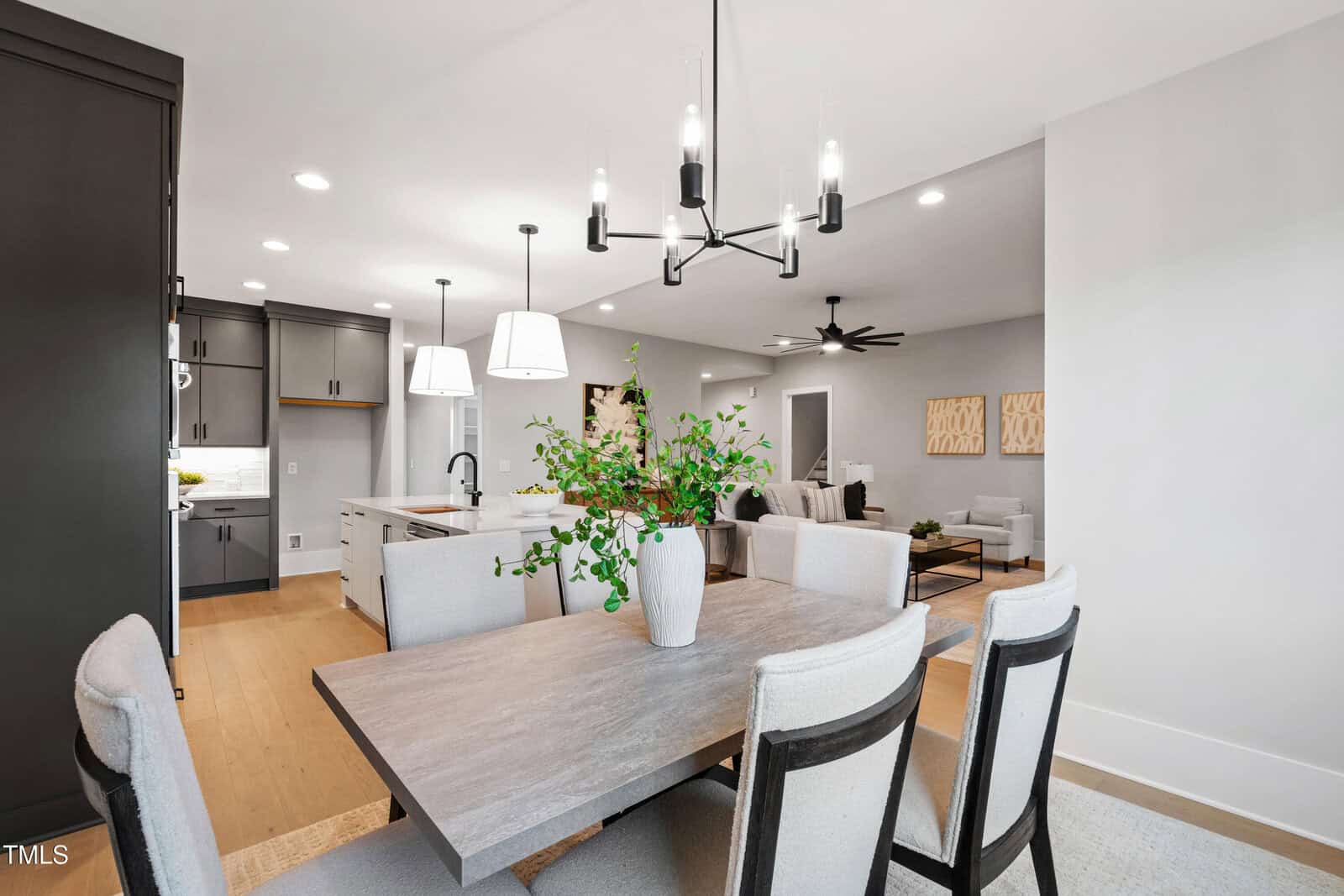 Modern open-concept kitchen and dining area with gray cabinets, wood floors, a light wood dining table, cushioned chairs, black chandeliers, and a plant centerpiece. Living room with sofas is visible in the background.
