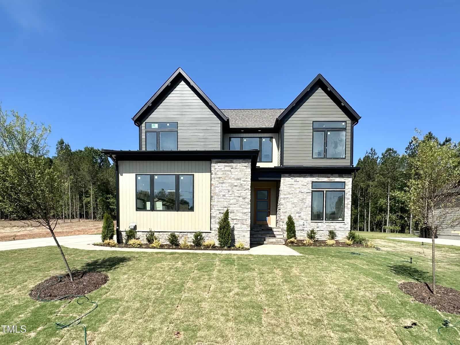 A two-story modern house with gray siding, stone accents, large windows, and a neatly landscaped front yard with small shrubs and young trees, set against a clear blue sky.