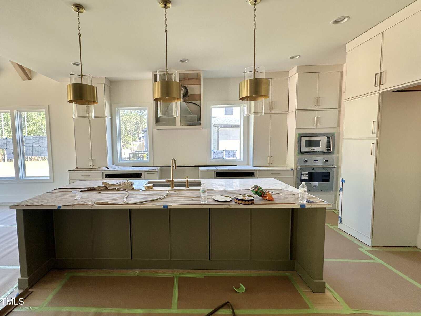 A modern kitchen with a large island covered in protective paper, brass pendant lights, built-in cabinets, an oven, a microwave, and two windows letting in natural light. The floor is taped off for protection.