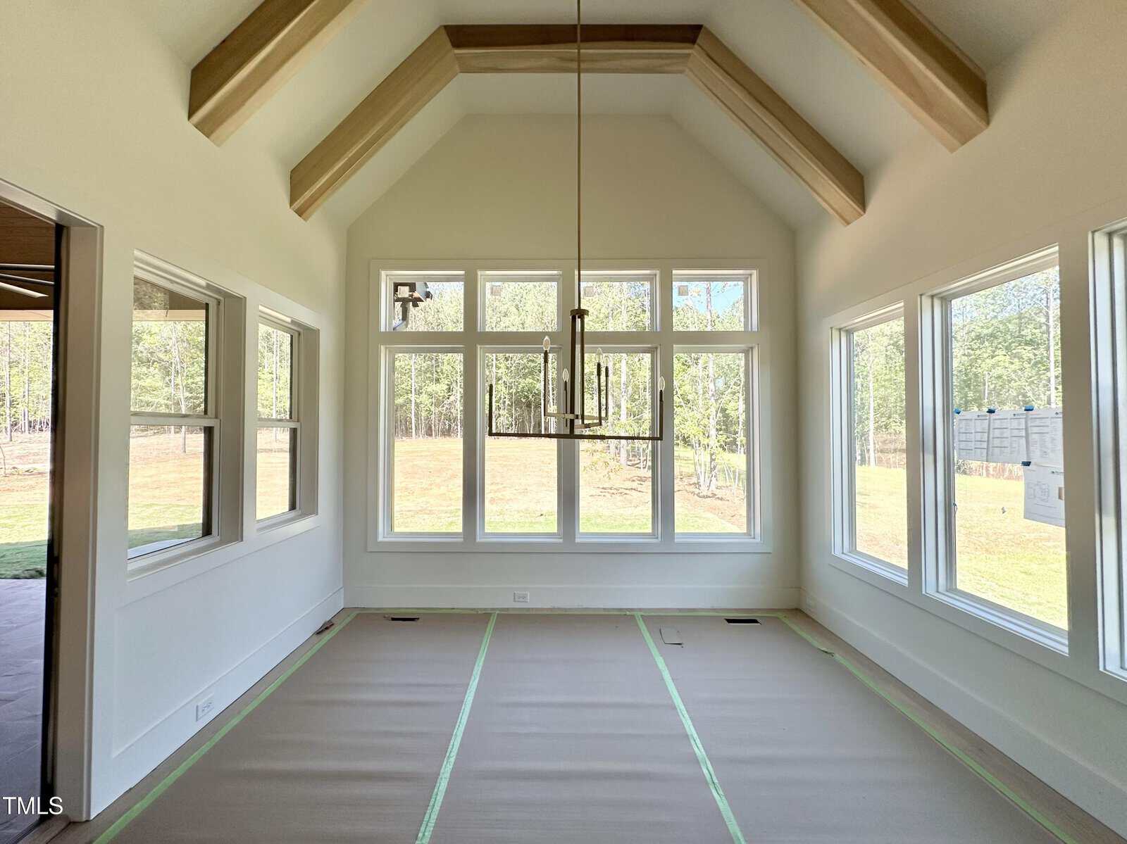 A bright, empty room with large windows on three sides, vaulted ceiling with exposed wooden beams, a modern chandelier, and protective covering on the floor with green tape along the edges.