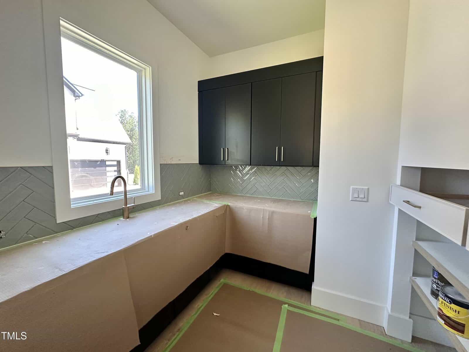 A modern kitchen under construction with dark upper cabinets, light backsplash in a herringbone pattern, a large window, and countertops covered with protective paper. Floors are also covered with protective material.