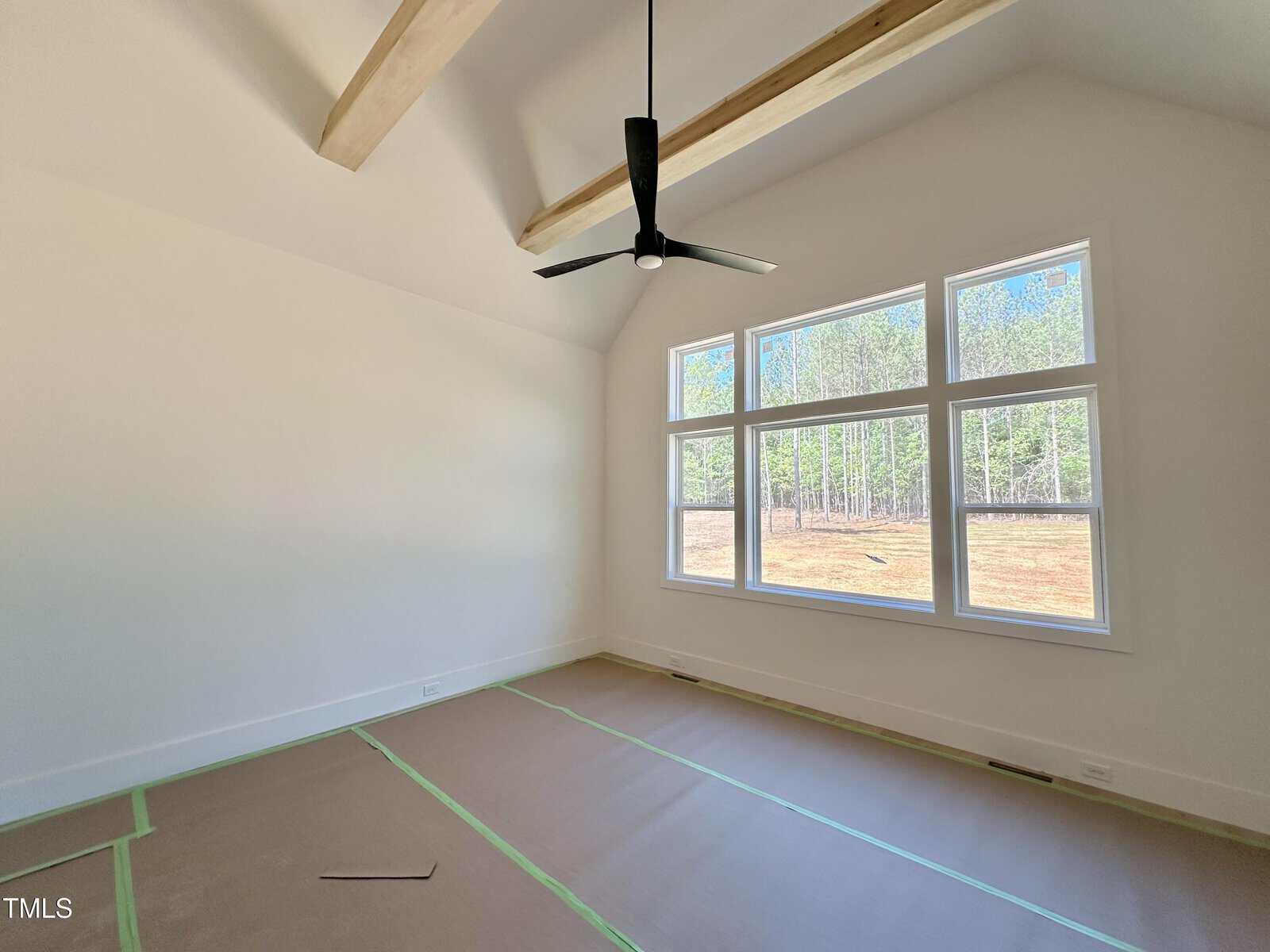 Bright, empty room with large windows, vaulted ceiling, exposed wooden beams, black ceiling fan, and floors covered in protective brown paper with green tape. Trees and grass are visible outside.