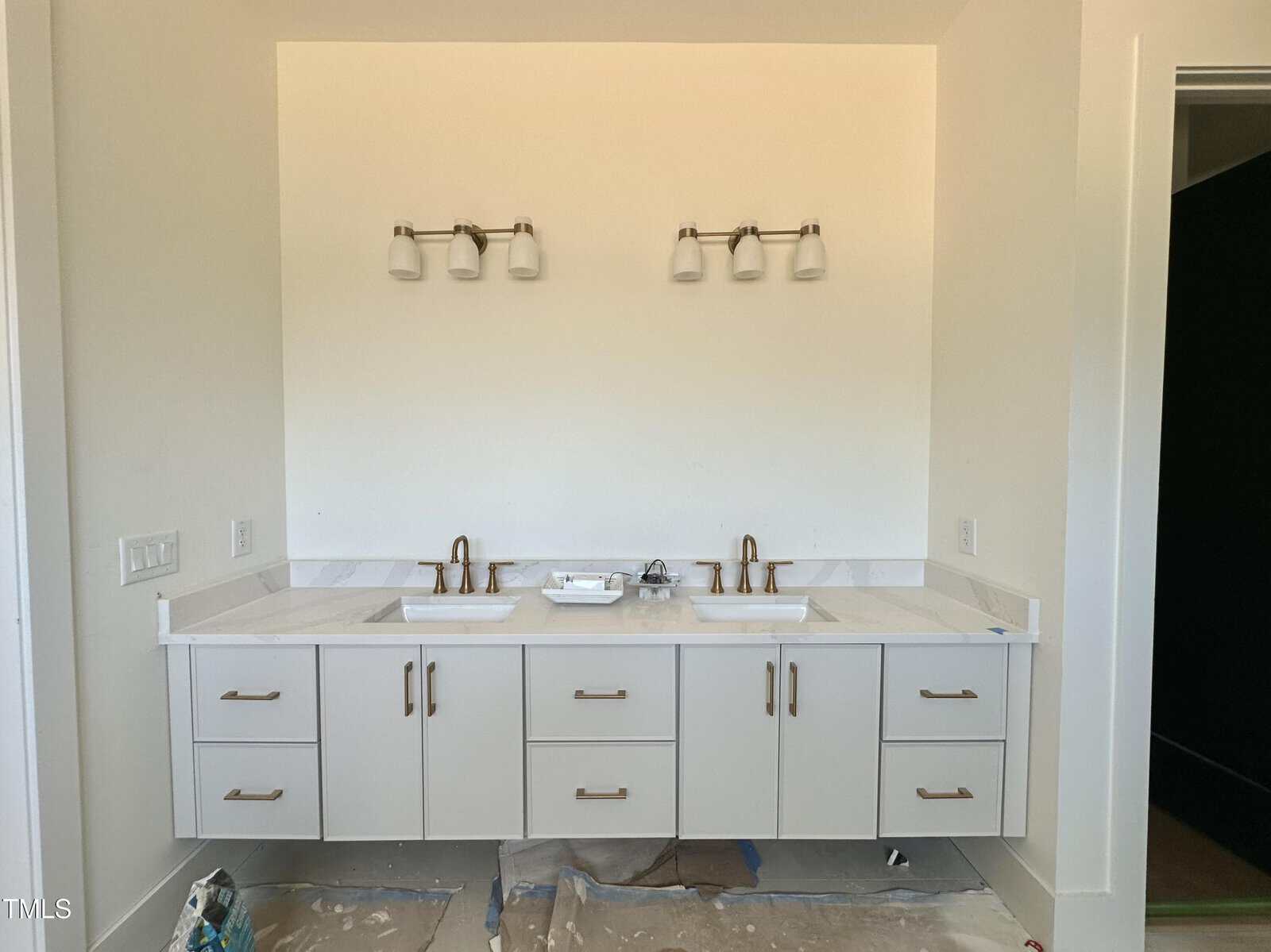 A modern double sink bathroom vanity with two mirrors missing, light fixtures above, white countertops, and light gray cabinets. The floor is unfinished with protective coverings and scattered materials.