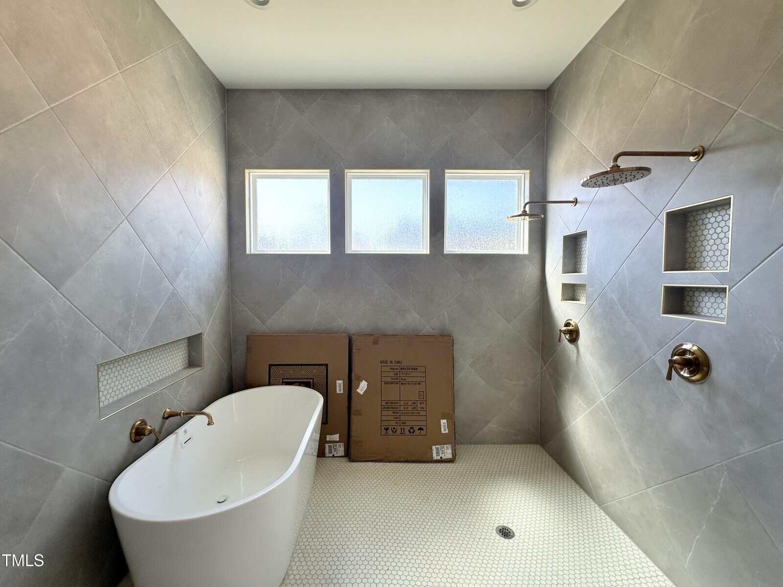 Modern bathroom with a freestanding tub, gray tiled walls, four small frosted windows, two showerheads, and built-in shelves. Cardboard boxes rest beside the tub. The floor is white hexagonal tile.