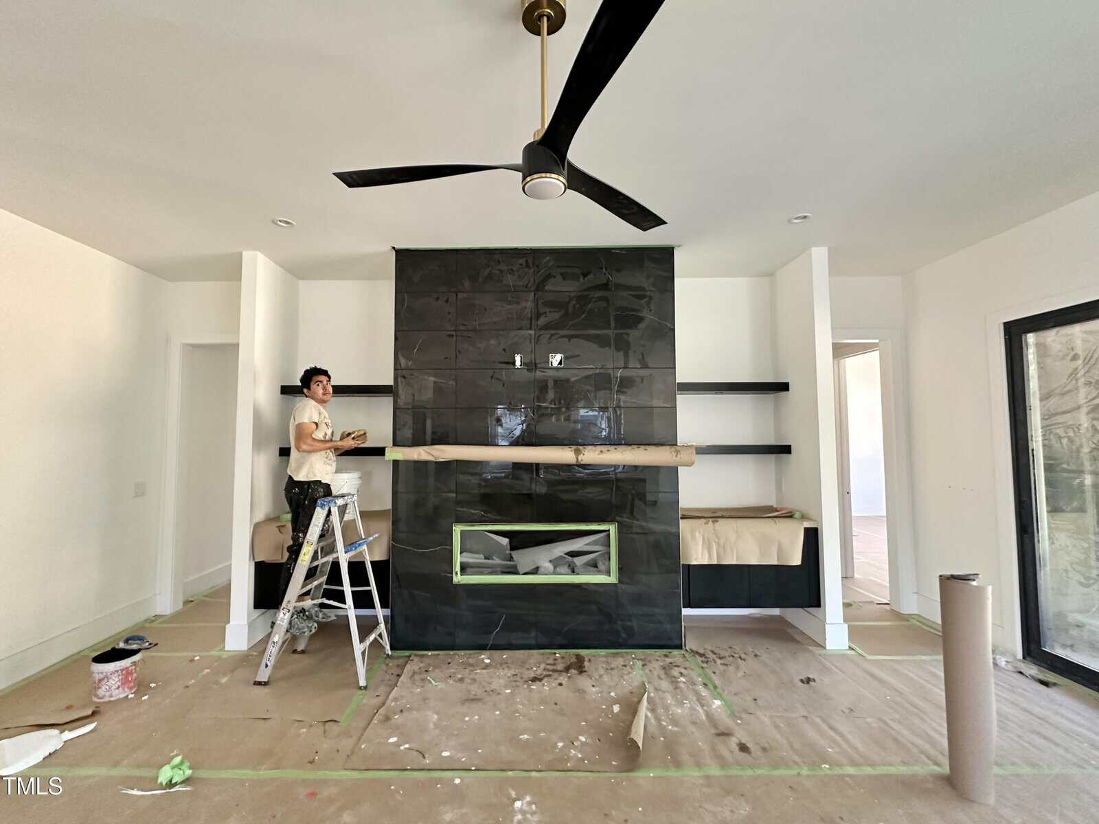 A person stands on a ladder working on a black tiled fireplace feature wall in a room under renovation. Shelves and floors are covered for protection, and a ceiling fan is visible above.