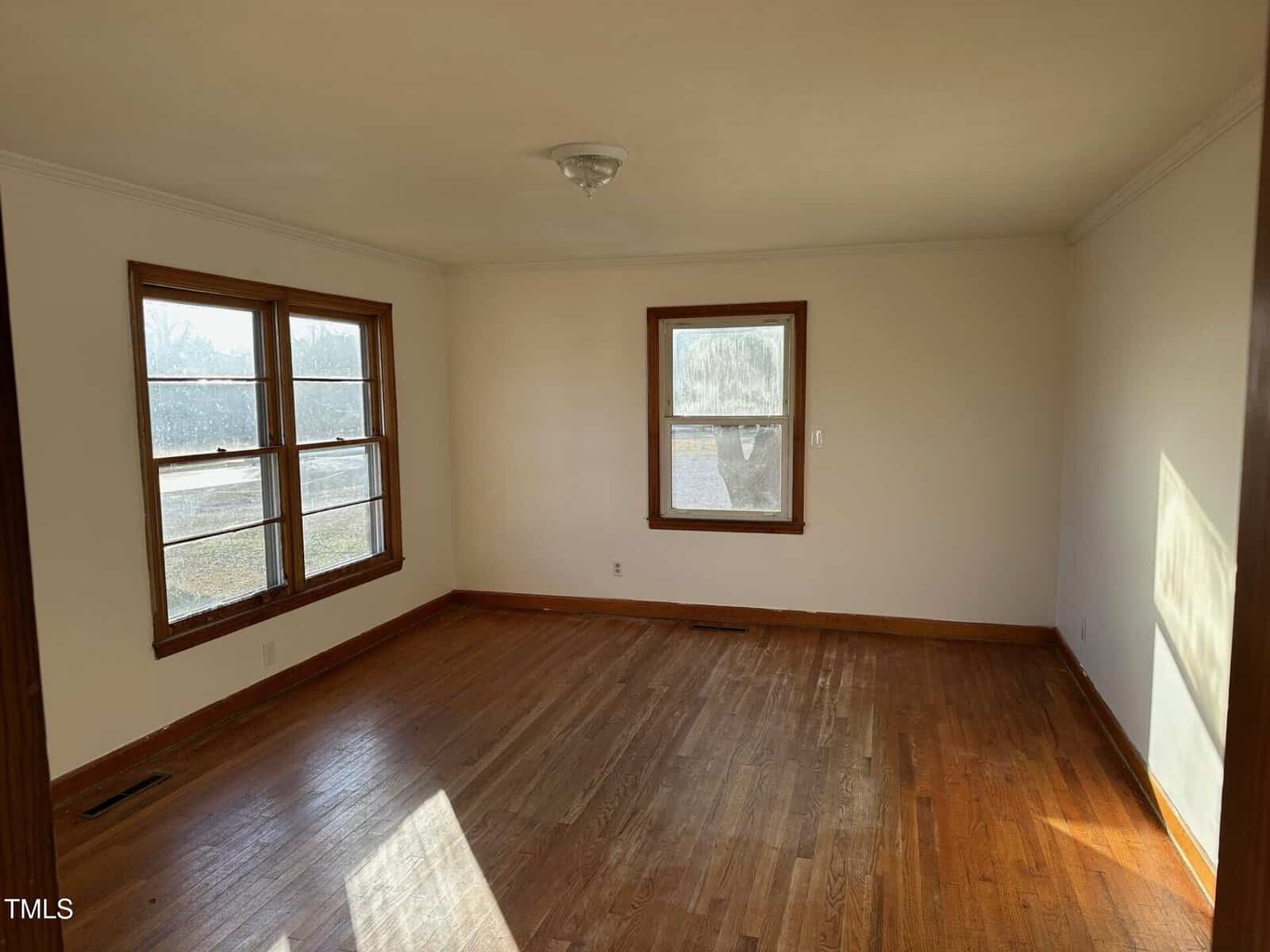 An empty room with hardwood floors and white walls. Two large windows on the left wall and one window on the back wall allow natural light to enter. A ceiling light fixture is centered on the ceiling.