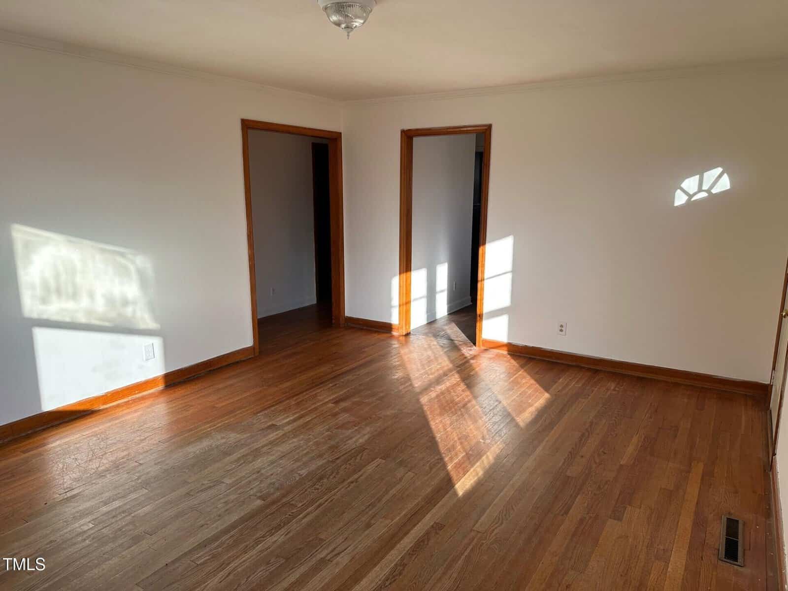 A vacant room with wooden floors and white walls. There are three doorways, each with wooden trim. Sunlight streams through a window, casting shadows on the floor and walls. A ceiling light fixture is visible.