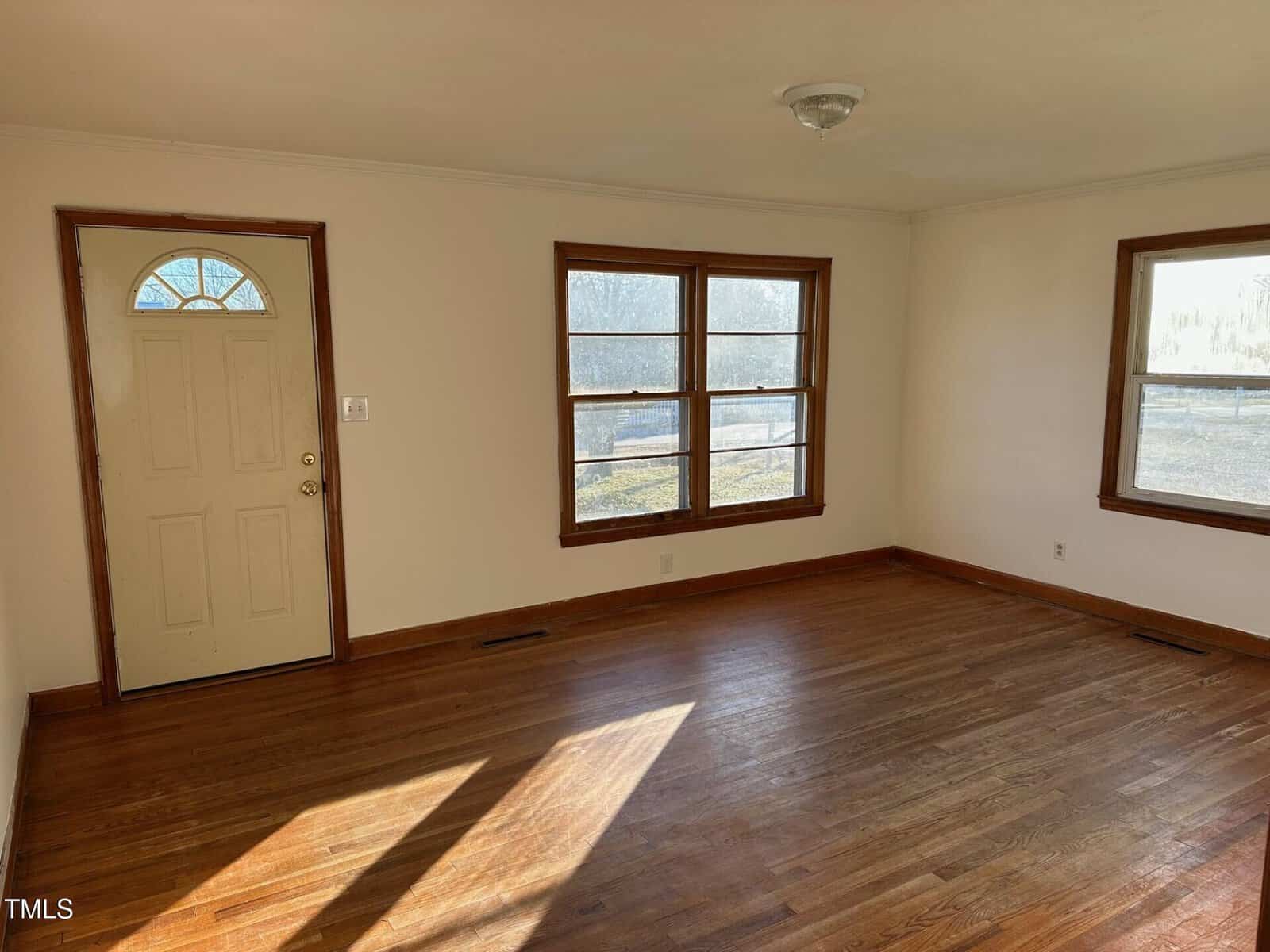 A small, empty living room with hardwood floors and white walls is shown. The room has two wooden-framed windows and a closed door with a window. Sunlight streams in, creating shadows on the floor. A ceiling light fixture is visible.