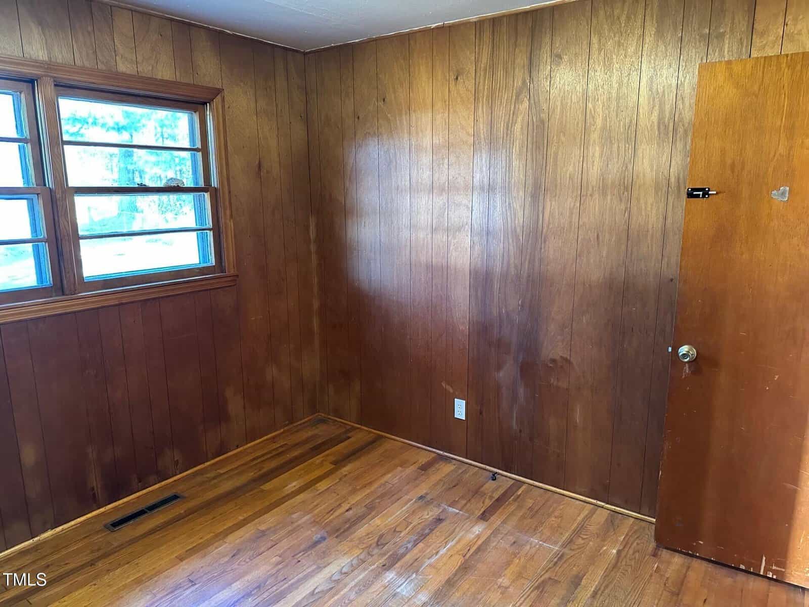 A small, empty room with wood-paneled walls and a wooden floor. There are two windows with horizontal panes on the left wall, and a closed wooden door on the right. A vent is visible in the bottom left corner of the room.