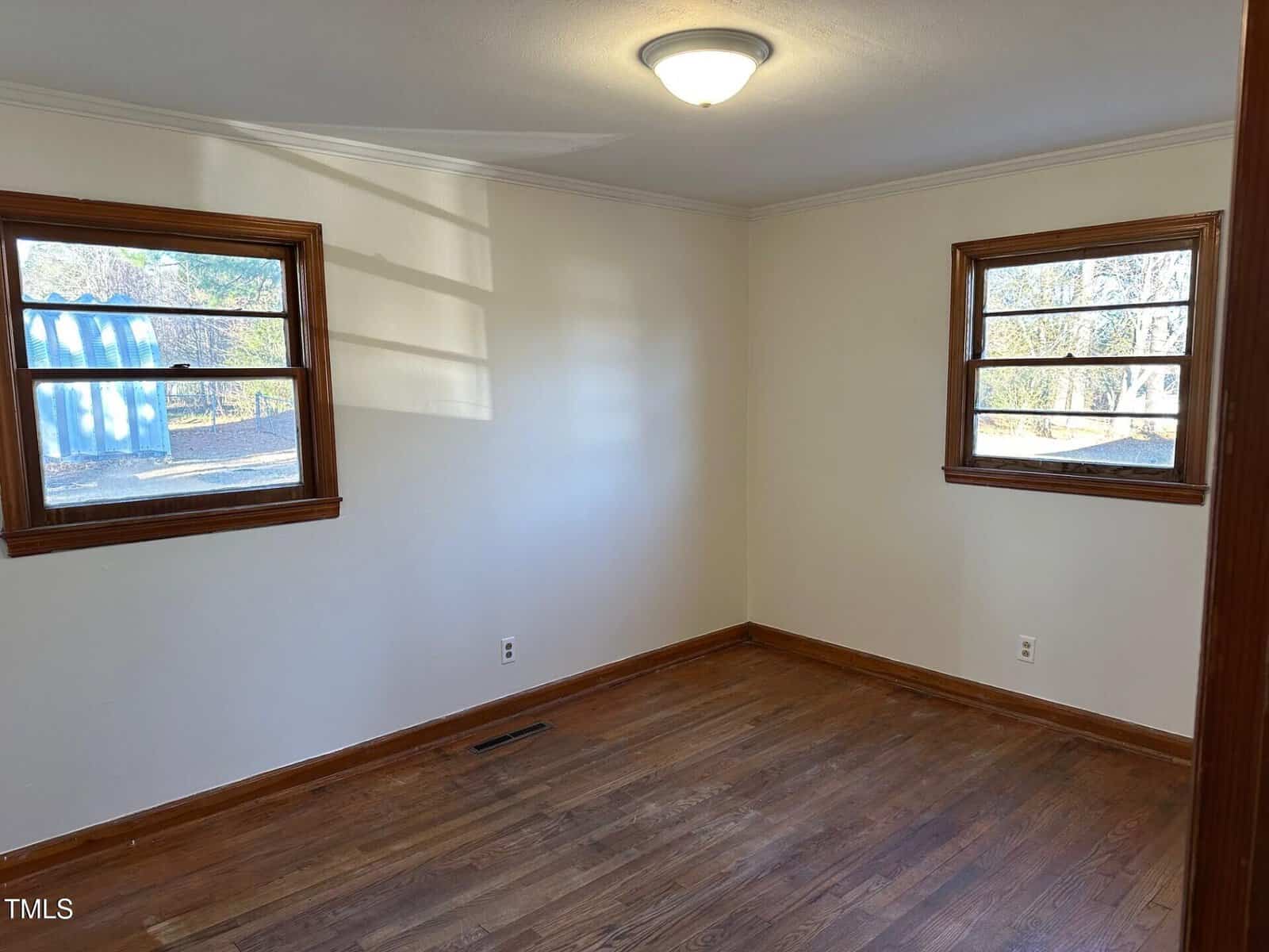 An empty room with white walls and hardwood flooring features two wooden-framed windows on adjacent walls. A ceiling light fixture is centered on the white ceiling. Sunlight streams in through the windows, casting shadows on the wall.