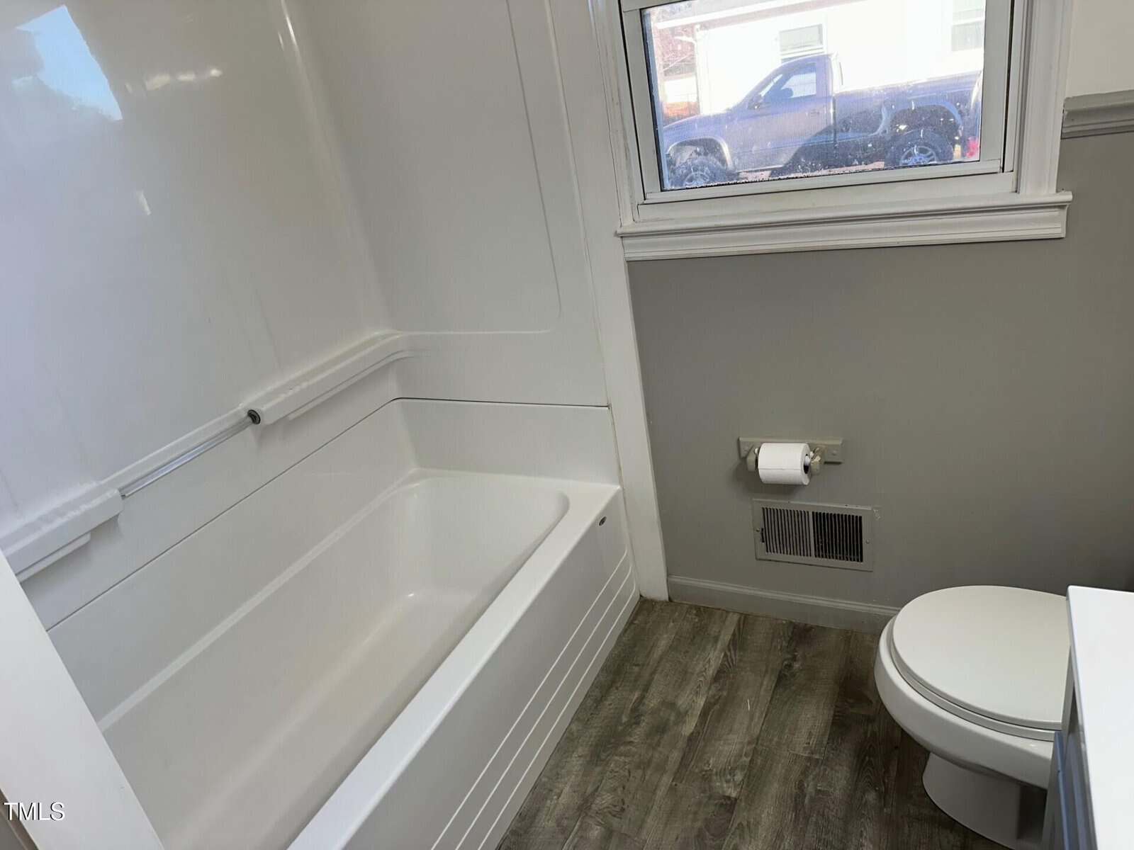 A bathroom with a white bathtub and a toilet on a wood-patterned floor. There is a wall-mounted toilet paper holder and a window showing a parked truck outside. The walls are light gray with white trim.