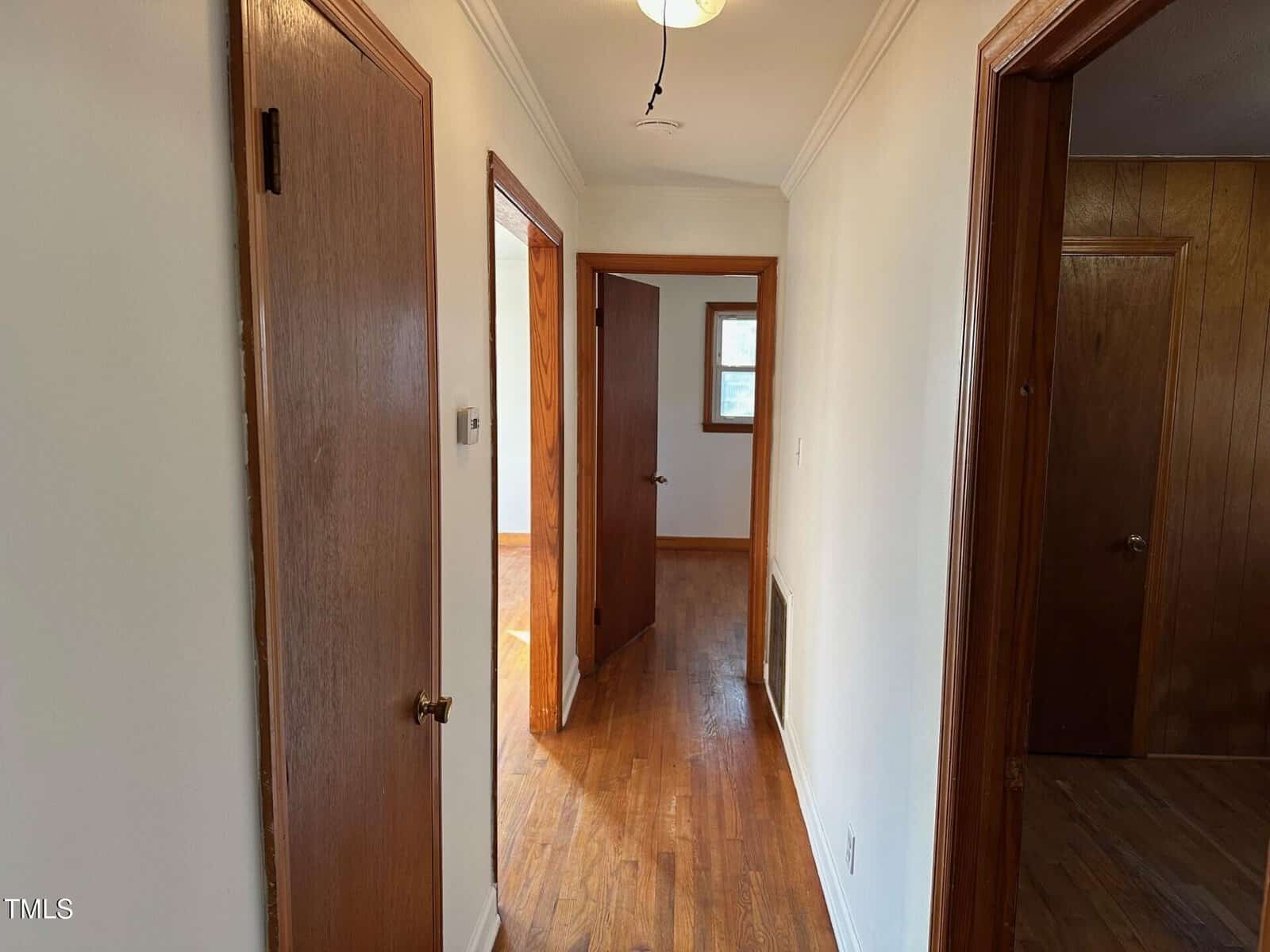 A hallway with hardwood floors and white walls, featuring three wooden doors on the left and right. A ceiling light fixture is visible. Sunlight filters through a small window at the end of the hallway.
