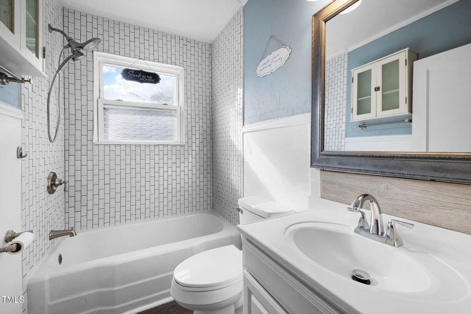 A bathroom features a white bathtub with a wall-mounted showerhead. The walls are tiled with a white rectangular pattern. A window is above the tub. There is a white sink with a mirror above it and a wall cabinet next to a toilet.