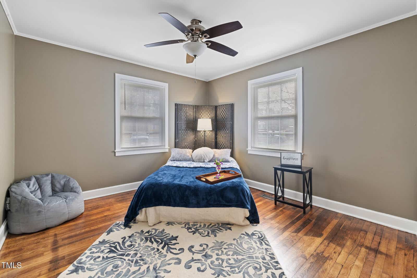 A bedroom with green walls features a double bed with a blue blanket and decorative pillow. There's a tray on the bed with a vase and book. The room has a ceiling fan, a small black side table, a beanbag chair, and a patterned rug on wooden floors.