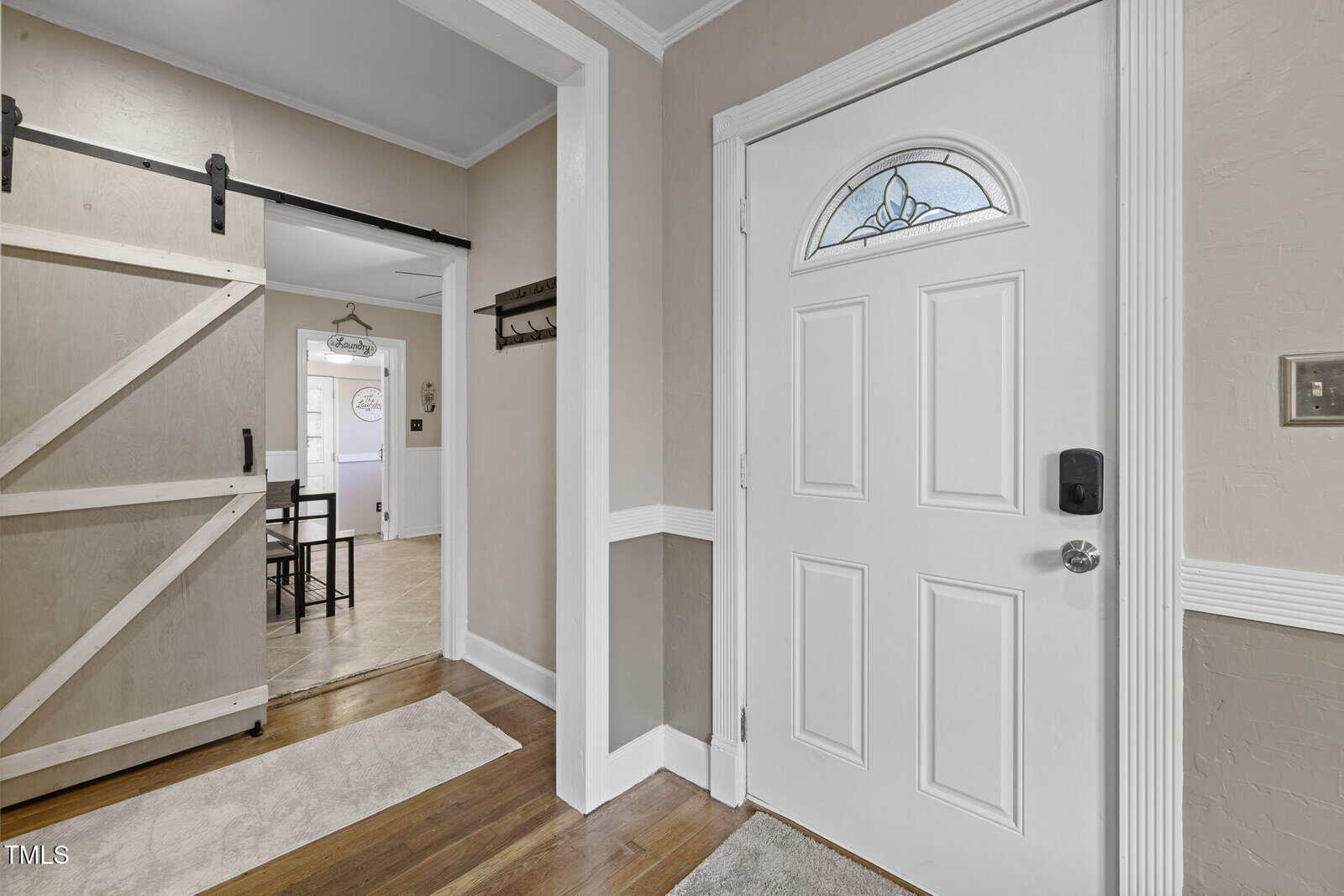 White door with an arched window section, next to a room with a sliding barn door. The wall has a white chair rail. A small rug covers part of the hardwood floor. A glimpse into a dining area with a table and chairs is visible.